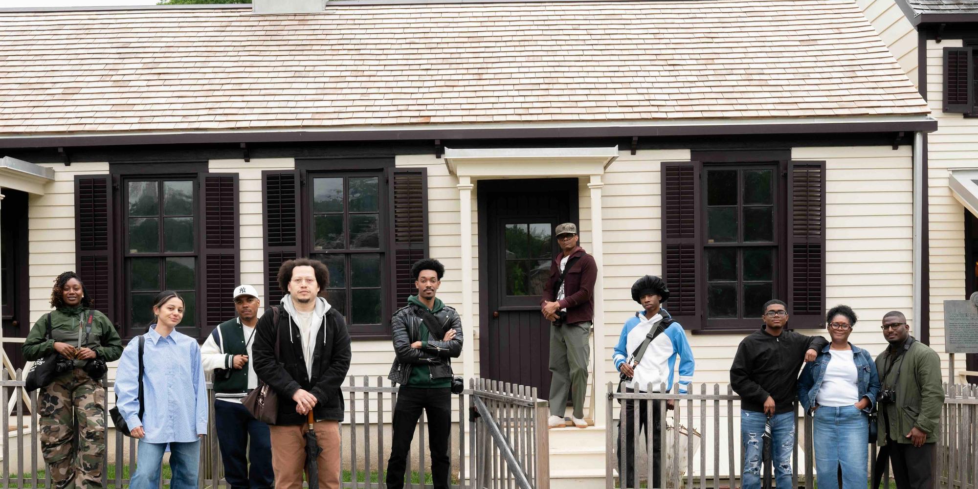A group of 10 people, a mix of men and women, are standing in front of a light-colored, clapboard building with a shingled roof. The building has dark shutters on its windows and a dark-colored door. A light-colored wooden fence runs horizontally across the foreground, with an open gate in the center. The ground is covered in light-colored gravel.
