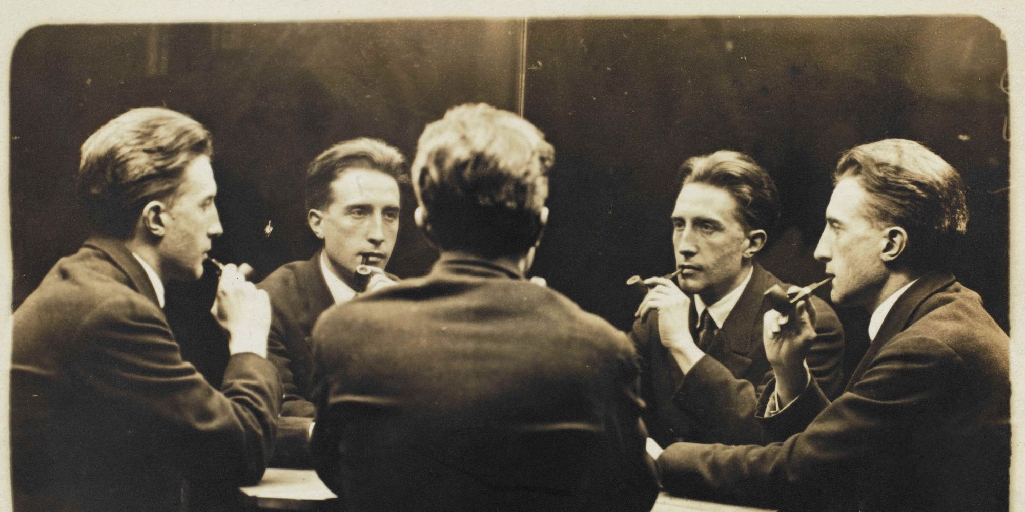 A man with slicked-back hair, smoking a pipe, sits at a table with four different versions of himself. 
