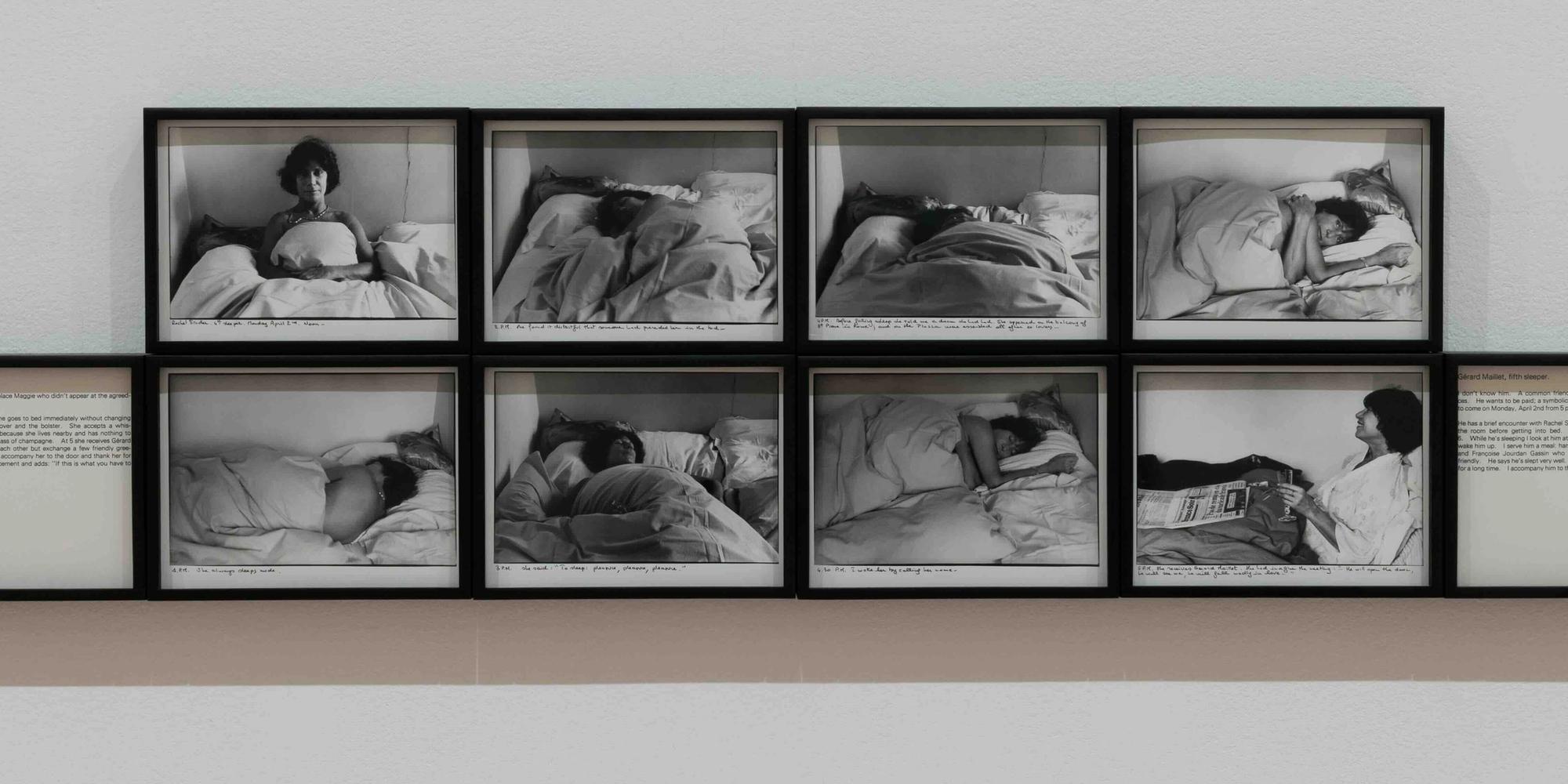 Large installation view showing a grid of many small black-and-white photographs arranged in rows on a wall, each depicting people sleeping or resting in bed